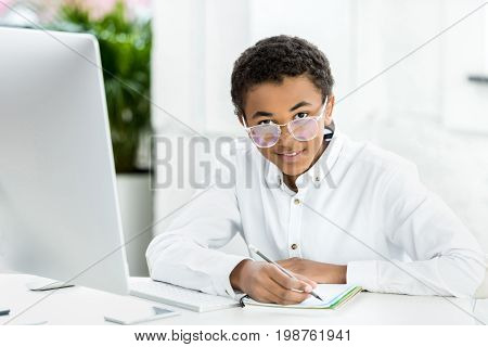portrait of smiling african american teenager in eyeglasses sitting at table and doing homework