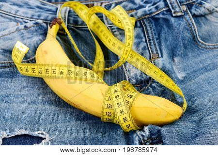 Banana wrapped with yellow measure tape on jeans selective focus. Mens denim pants with banana imitating male genitals. Health and male sexuality concept. Jeans loops zipper and pocket close up.