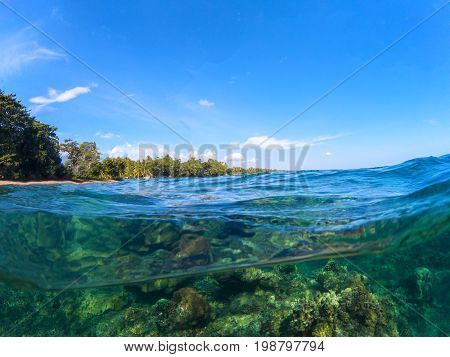 Split landscape with sea and sky. Split photo with tropical island and underwater coral reef. Clear blue sky and seawater. Above and below waterline sea view. Exotic seashore panorama. Split seascape