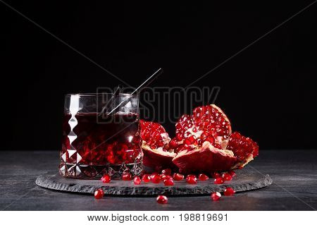 A transparent glass of healthy pomegranate juice and a cut garnet on a round plate on a saturated black background. Organic summer fruity red cocktail with straws and fresh garnet.