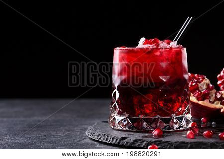 Close-up of a transparent glass of sweet pomegranate juice with ice and a cut garnet on a round plate on a saturated black background. Healthful and fruity red cocktail with straws and fresh garnet.