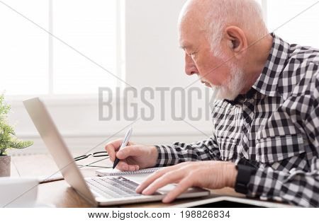 Mature man using laptop and writing in notepad at home desk. Education, recipe concept