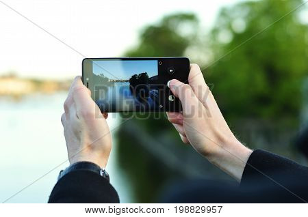 Photo of a man's hands holding phone and taking photo of a river with blurred trees and water in the background