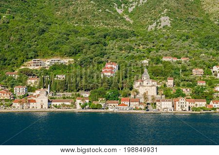 Prcanj - a small town along the Bay of Kotor Montenegro. A part of the waterfront that consists of a long line of stone villas and churches separated by gardens and olive orchards with the baroque Birth of Our Lady church to the right.