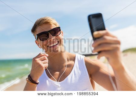 summer holidays and people concept - happy smiling young man with earphones and smartphone taking selfie on beach