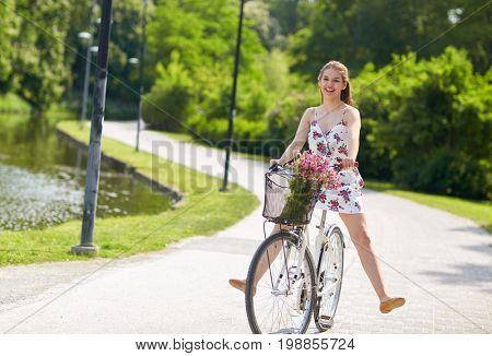 people, leisure and lifestyle - happy young hipster woman wearing summer dress riding fixie bicycle with wild flowers in basket at park