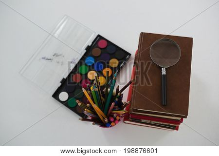 Overhead view of book stack, magnifying glass, color pencils and palette on white background