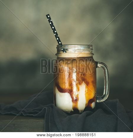Iced caramel macciato coffee with milk in glass jar with straw on dark rustic wooden table, selective focus, copy space, square crop. Cold sweet refreshing summer drink concept