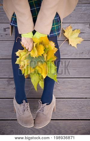 Top view on woman's hands holding beautiful bunch of yellow marple leaves. Young woman with bouquet of autumn leaves sitting on wooden bridge in park.autumn thanksgivining and lifestyle concept
