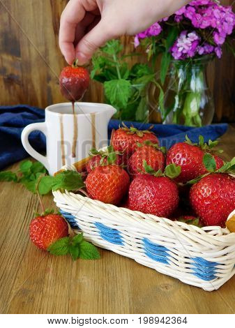 Strawberries in a wicker basket and chocolate sauce in a ceramic jug. Female hand is dipping strawberry into the sauce