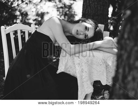 Portrait Of A Young Woman In A Long Evening Dress, She Sits At A Table In The Woods. Black And White