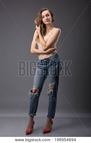Studio portrait of pretty woman model with amazing body long legs leaning agains wall wearing denim jeans lingerie top high heels holding jacker looking at camera. Over gray background