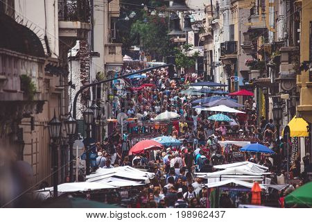 The market is in the street in Argentina. The narrow street of Buenos Aires is crowded with people. The street is decorated with street umbrellas. The foreground is blurred.