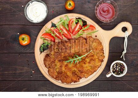 A large Viennese schnitzel and tomato salad on a cutting board on a dark wooden background. Top view.