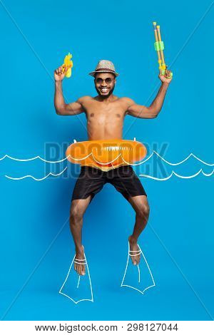 Summer Vacation Concept. Happy African American Guy In Swimwear And Inflatable Ring Jumping With Wat