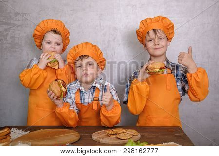 Three Cute Boys In Costumes Cooks Engaged In Cooking Homemade Burgers. Three Brothers Preparing Fami