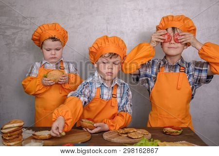 Three Cute Boys In Costumes Cooks Engaged In Cooking Homemade Burgers. Three Brothers Preparing Fami