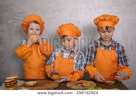 Three Cute Boys In Costumes Cooks Engaged In Cooking Homemade Burgers. Three Brothers Preparing Fami