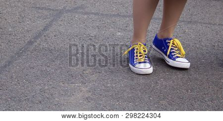 Ankles Of Girl Wearing Blue Sports Shoes With Yellow Laces Against Grey Pavement Background - Image