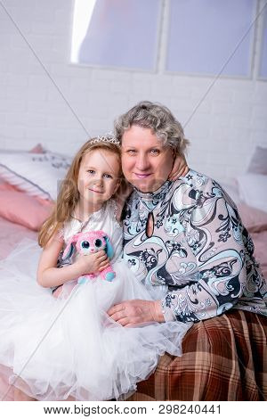 Happy Little Girl And Her Grandmother Are Sitting Together And Hugging In The Bedroom. They Are Smil