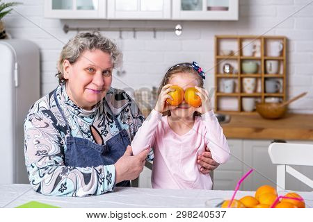 Happy Little Girl And Her Grandmother Have Breakfast Together In A White Kitchen. They Are Having Fu