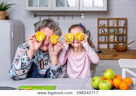 Happy Little Girl And Her Grandmother Have Breakfast Together In A White Kitchen. They Are Having Fu