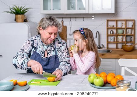 Happy Little Girl And Her Grandmother Have Breakfast Together In A White Kitchen. They Are Having Fu