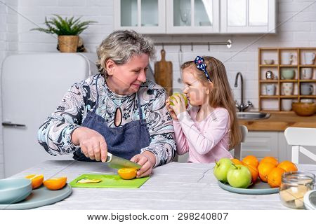 Happy Little Girl And Her Grandmother Have Breakfast Together In A White Kitchen. They Are Having Fu