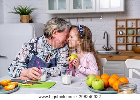 Happy Little Girl And Her Grandmother Have Breakfast Together In A White Kitchen. They Are Having Fu