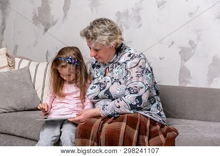 Grandmother And Her Little Granddaughter Are Watching Movies Together And Playing On The Device Whil