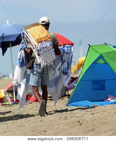 Street Vendor Walks On The Beach To Sell The Many Rugs He Carries On His Shoulder