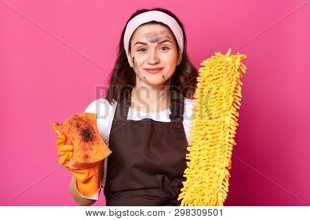 Slender Adorable Attractive Girl Looks Directly At Camera With Bright Broom And Sponge In Both Hands