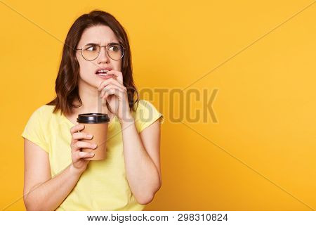 Confused Thoughtful Young Lady Looks Aside, Puts Finger In Her Mouth, Holds Papercup With Coffee In 