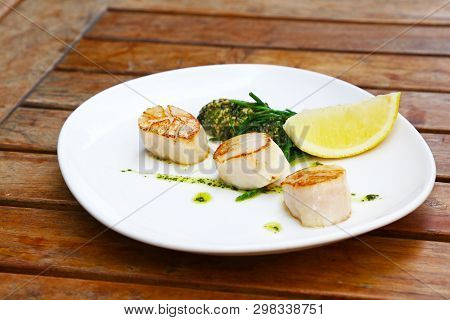 Close Up Portion Of Three Grilled Scallops On White Plate Over Brown Wooden Table, High Angle View