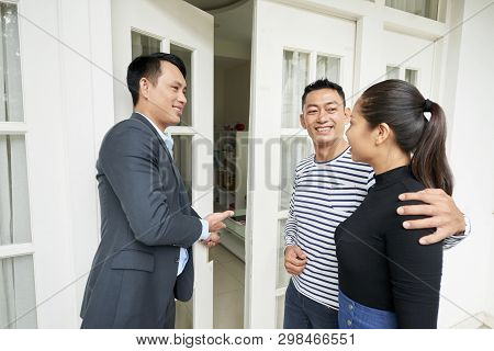 Asian Real Estate Agent Opening The Door Of The House And Inviting Young Couple To Come Inside And S