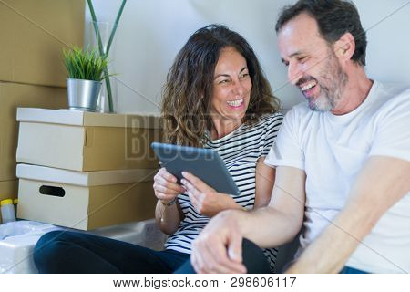 Middle age senior romantic couple in love sitting on the apartment floor with boxes around and using touchpad tablet smiling happy for moving to a new home