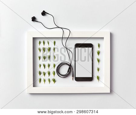 Creative ogranic herbal pattern of pine needles twigs, mobile smartphone with blank screen for mockup and headphones in a frame on a light gray background. Place for text. Flat lay.