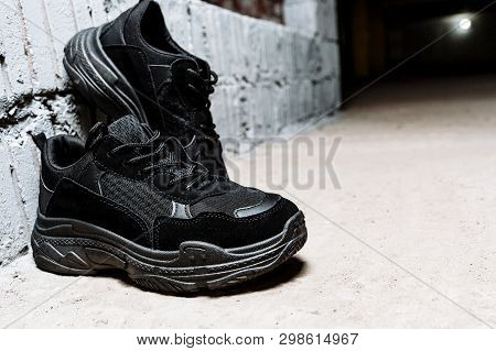 Pair Of Black Sneakers With Thick Soles On The Cement Floor Against The Background Of White Wall