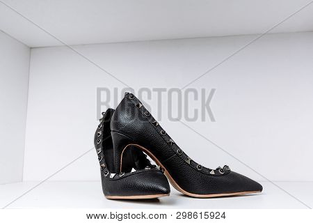 Pair Of Black High-heeled Shoes With Pointed Toes, Decorated With Metal Inserts Against A Shelf In T