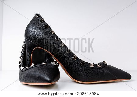 Pair Of Black High-heeled Shoes With Pointed Toes, Decorated With Metal Inserts Against A Shelf In T