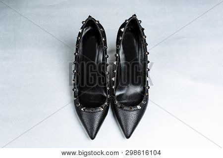 Pair Of Black High-heeled Shoes With Pointed Toes, Decorated With Metal Inserts Against A Shelf In T
