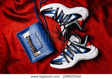White Sneakers With Black And Blue Inserts On A Thick Figured Sole And A Blue Bag With A Gold Chain