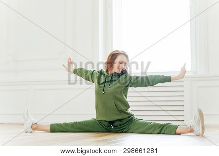 Indoor Shot Of Fit Female Stretches Hand And Legs Sideways, Sits In Pose Of Splits, Tilts Head On Ri