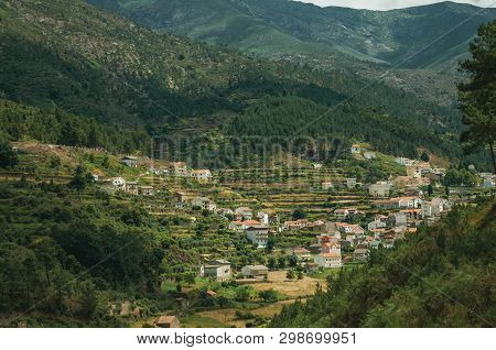 Hilly Landscape Covered By Trees And Terraced Fields With The Roofs Of Alvoco Da Serra Underneath. A