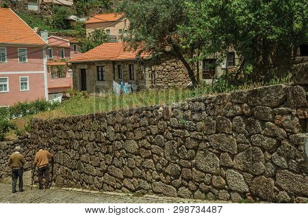 Alvoco Da Serra, Portugal - July 15, 2018. Two Old People Walking Down The Alley On Slope Next To St