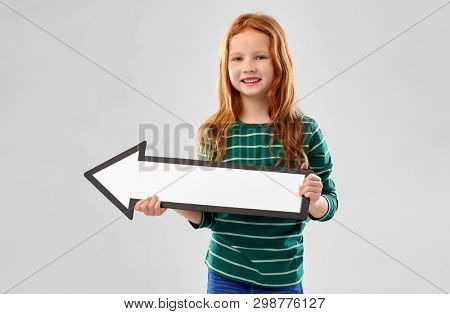 childhood, fashion and people concept - smiling red haired girl in green striped shirt holding big white leftwards thick arrow over grey background
