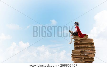 Little Child In Mask And Cape Sittting On Pile Of Old Books In Sky And Reading