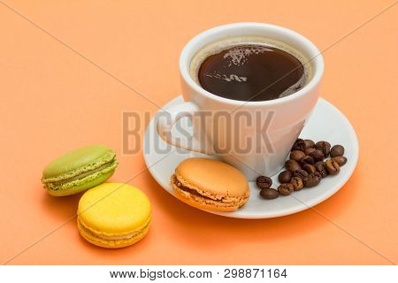 White Cup Of Coffee With Coffee Beans And Delicious Macarons Cakes Of Different Color With Peach Col