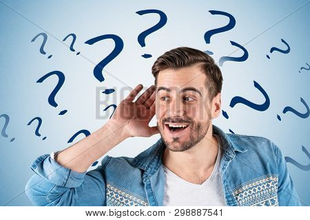 Portrait Of Smiling Young Man In Casual Clothes Listening Attentively With His Palm Near Ear Standin