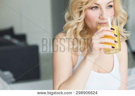 Attractive Blonde Woman Sits On The Edge Of The Bed At Home And Holds A Stocker With Orange Juice In